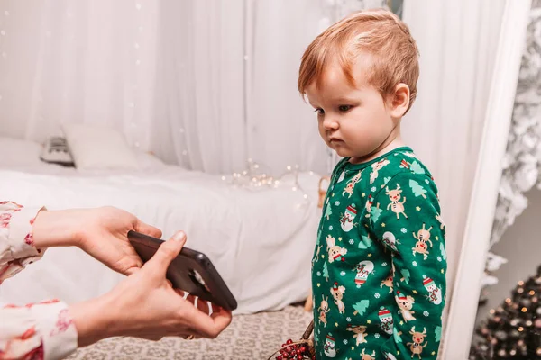 Happy cheerful young small kid dressed green warm pajama standing and playing with phone, celebrating New Year or Christmas at home. Boy preparing for sleeping night, addiction to gadgets