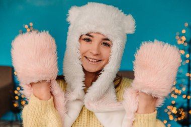 Sıcak süveter ve yün şapka giyip poz vererek kutlama yapan genç ve güzel bir kadın. Kış tatili panayırı, seyahat ruhları beldesi. Yılbaşı süsleri, çelenkler