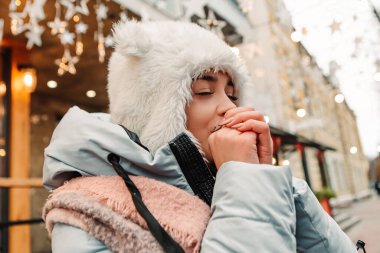 Gülümseyen kadın sıcak ceket ve yün şapkayla ellerini ağızdan buharla ısıtarak kutluyor. Kış tatili panayırı, seyahat ruhları beldesi. Yılbaşı süsleri, çelenkler