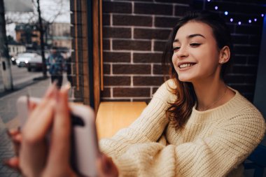 Sıcak kazak giymiş gülümseyen bir kadın, telefon aleti tutan ve sokak kafesinde selfie çeken bir kadın. Bayan arkadaşlarıyla sohbet ediyor, video görüşmesi yapıyor, konuşuyor.