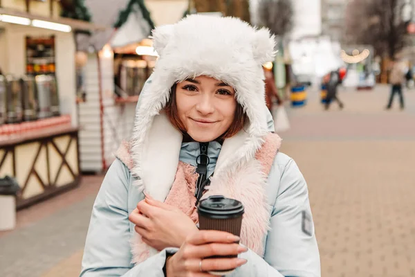暖かいジャケットとウールの帽子を身に着けていることを祝う若いかなり笑顔の女性は ホットティーコーヒー飲料マルチワインを飲みます 冬のマーケットの休日フェア 新年のクリスマスの装飾 ガーランド — ストック写真