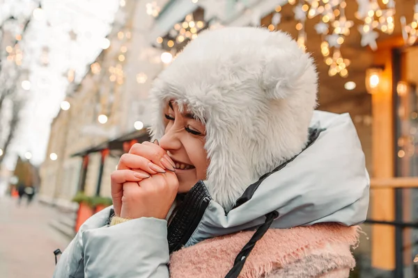 笑着庆祝的女人穿着暖和的夹克和羊毛帽子 用蒸汽从嘴里加热她的手 冬季市场假期展会 旅游精神度假胜地 新年装饰品 — 图库照片