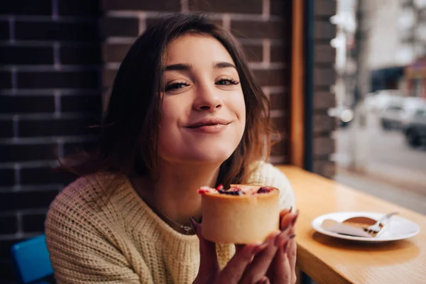 stock image Smiling woman dressed warm sweater, enjoy eating dessert, tasty cupcake. Snack food in street cafe. New Year Christmas decorations, garlands