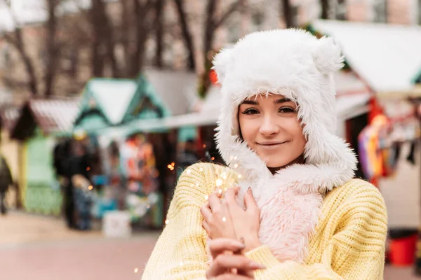 暖かいセーターと織られたウールの帽子を着て祝う若いかなり笑顔の女性は 優しい光の輝きを保持します 冬の市場の休日フェア 旅行精神リゾート 新年のクリスマス飾り — ストック写真