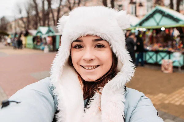 若いかなり笑顔の女性は 携帯電話でシーリングフィーを取り 暖かいジャケットと織られたウールの帽子を着て祝う 冬の市場の休日フェア 旅行精神リゾート 新年のクリスマス飾り — ストック写真