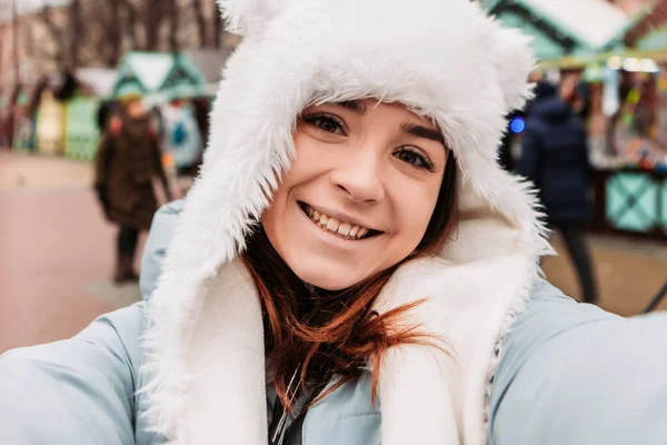 若いかなり笑顔の女性は 携帯電話でシーリングフィーを取り 暖かいジャケットと織られたウールの帽子を着て祝う 冬の市場の休日フェア 旅行精神リゾート 新年のクリスマス飾り — ストック写真