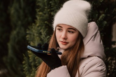 Genç, güzel bir kadın, kalın bir ceket giymiş, Noel Ağacı 'nın yanında duruyor, telefonla konuşuyor, sesli mesaj yolluyor. Kış tatili, tatil beldesi. Yeni yıl Noel süslemeleri