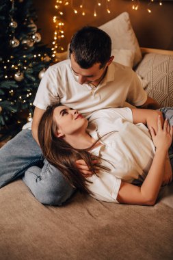 Dikey, genç, romantik bir çift yatak çelenklerinde uzanıyor. Evlerine sarılıyorlar. Yeni yıl ağacı süslemeleri. 