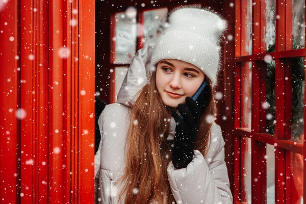 若いかなり女性の暖かいジャケットを着て 電話ボックスの近くに立って 電話をかけ 雪が降っている 冬の市場の休日フェア 旅行精神リゾート 新年のクリスマス飾り — ストック写真