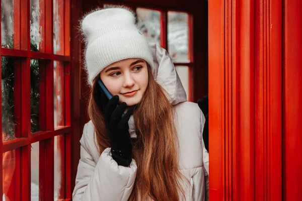 Young pretty smiling woman dressed warm jacket, standing near telephone red box, talking phone. Winter market holidays fair, travel spirit resort. New Year Christmas decorations