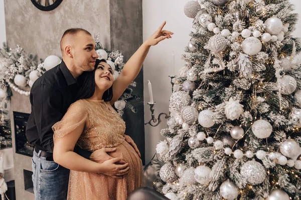 男人抱着一个肚子圆圆的女人 把小孩抱在里面装饰圣诞树 庆祝新年 怀孕的女士在等婴儿 怀孕的新生儿 — 图库照片