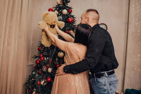 男人抱着一个肚子圆圆的女人 带着小孩在圣诞树内装饰 抱着泰迪熊 庆祝新年 怀孕的女士在等婴儿 怀孕的新生儿 — 图库照片