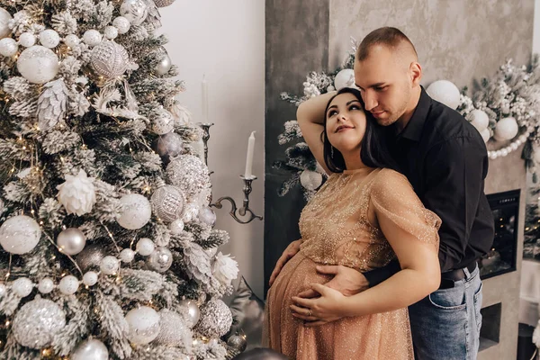 男人抱着一个肚子圆圆的女人 把小孩抱在里面装饰圣诞树 庆祝新年 怀孕的女士在等婴儿 怀孕的新生儿 — 图库照片