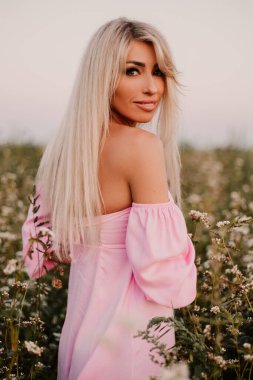 Vertical photo blonde woman posing in the big endless field of daisies in summer evening. Lady wear pink dress, looking at the camera, touching the plant green blossoming flower. Moody atmosphere