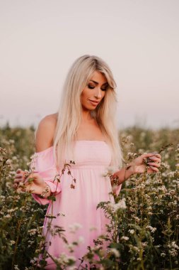 Vertical photo blonde woman posing in the big endless field of daisies in summer evening. Lady wear pink dressed, looking aside smiling, touching the plant green blossoming flower. Moody atmosphere