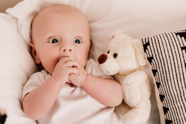 Little boy funny cute child baby lying on bed pillows with fluffy toy. Playful toddler with bulging eyes having fun, making faces grimaces. Happy childhood, family concept