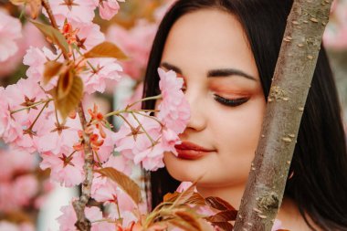 Portrait of beautiful tender woman dressed pink flowery dress posing near sakura cherry blossoms blooming flowers in the garden park in early spring. Hanami celebration, Japanese festival