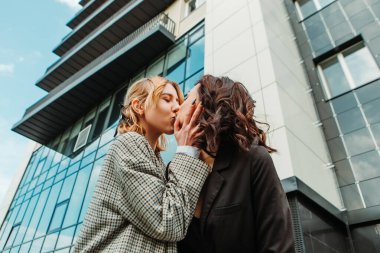 Two pretty women friends posing near glass building. Couple of gay lesbian girls hugging embracing kissing girlfriends, dressed casual outfits, have a date. LGBT concept. Fashion, make up, hairstyle