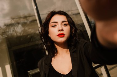 Pretty woman posing near glass building. Girl dressed casual outfits makes selfie, holding camera phone. Fashion, make up, hairstyle