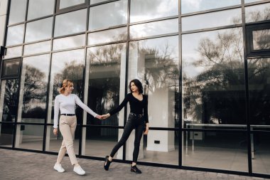 Two pretty women friends posing near glass building. Couple of gay lesbian girls hugging embracing walking girlfriends, dressed casual outfits, have a date. LGBT concept. Fashion, make up, hairstyle