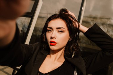 Pretty woman posing near glass building. Girl dressed casual outfits makes selfie, holding camera phone. Fashion, make up, hairstyle