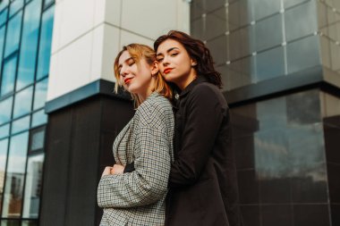 Two pretty women friends posing near glass building. Couple of gay lesbian girls hugging embracing together girlfriends, dressed casual outfits, have a date. LGBT concept. Fashion, make up, hairstyle