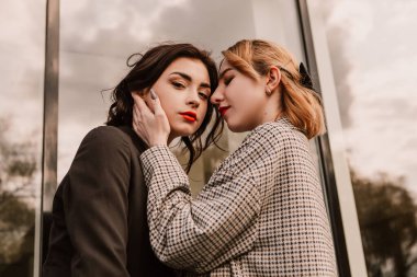 Two pretty women friends posing near glass building. Couple of gay lesbian girls hugging embracing kissing girlfriends, dressed casual outfits, have a date. LGBT concept. Fashion, make up, hairstyle