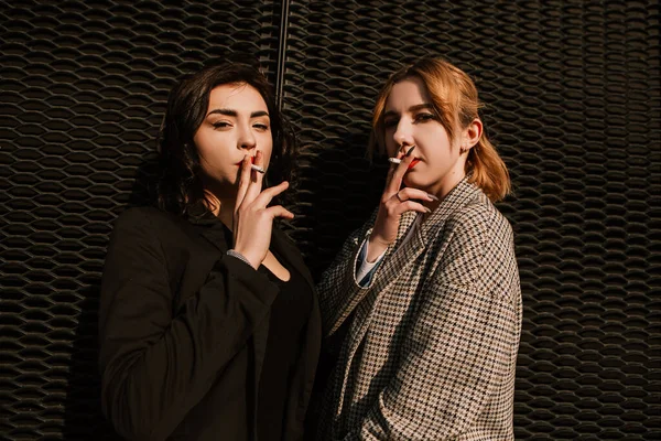 Two pretty women friends standing outside smoking cigarette. Couple of gay lesbian girls hugging embracing together girlfriends, have a date. LGBT concept. Addiction, cancer, tobacco. Bad habit