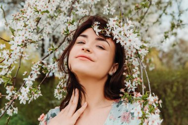 Portrait of charming pretty woman dressed flowery dress posing near apple cherry tree blossoms blooming flowers in the garden park in early spring nature. Fashion, girl model with black hair