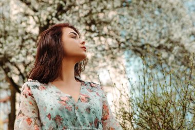 Portrait of charming pretty woman dressed flowery dress posing near apple cherry tree blossoms blooming flowers in the garden park in early spring nature. Fashion, girl model with black hair