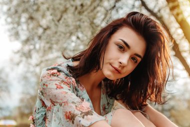 Portrait of charming pretty woman dressed flowery dress sitting on bench near apple cherry tree blossoms blooming flowers in the garden park in early spring nature. Fashion, holiday