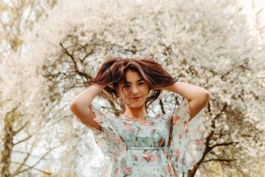 Portrait of charming pretty woman dressed flowery dress posing touching hair near apple cherry tree blossoms blooming flowers in the garden park in early spring nature. Fashion holiday