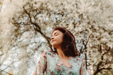 Portrait of charming pretty woman dressed flowery dress posing near apple cherry tree blossoms blooming flowers in the garden park in early spring nature. Fashion, girl model with black hair