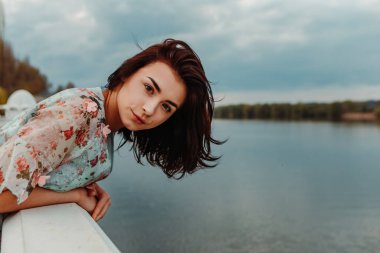 harming pretty woman dressed flowery dress standing on the pier near river lake moody cloudy weather in early spring nature. Fashion, girl model with black hair
