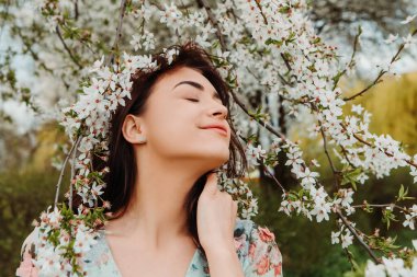 Portrait of charming pretty woman dressed flowery dress posing near apple cherry tree blossoms blooming flowers in the garden park in early spring nature. Fashion, girl model with black hair