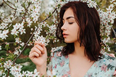 Profile pose looking to side woman dressed flowery dress posing near apple cherry tree blossoms blooming flowers in the garden park in early spring nature. Fashion, girl model with black hair