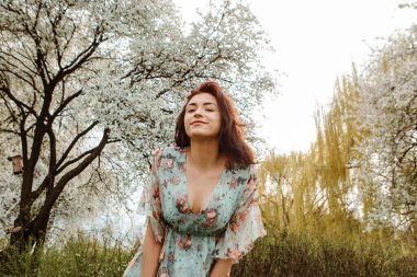 Portrait of charming pretty woman dressed flowery dress posing near apple cherry tree blossoms blooming flowers in the garden park in early spring nature. Fashion, girl model with black hair