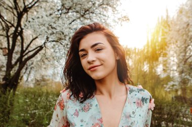 Portrait of charming pretty woman dressed flowery dress posing near apple cherry tree blossoms blooming flowers in the garden park in early spring nature. Fashion, girl model with black hair