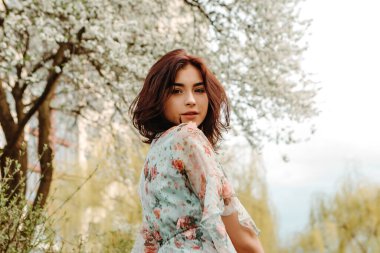 Woman looking over the shoulder dressed flowery dress posing near apple cherry tree blossoms blooming flowers in the garden park in early spring nature. Fashion, girl model with black hair