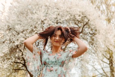 Portrait of charming pretty woman dressed flowery dress posing touching hair near apple cherry tree blossoms blooming flowers in the garden park in early spring nature. Fashion holiday
