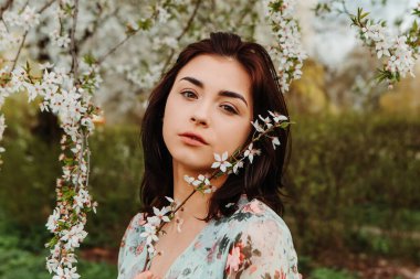 Portrait of charming pretty woman dressed flowery dress posing near apple cherry tree blossoms blooming flowers in the garden park in early spring nature. Fashion, girl model with black hair