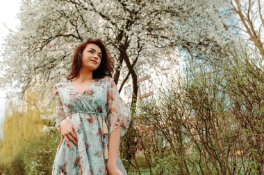 Portrait of charming pretty woman dressed flowery dress posing near apple cherry tree blossoms blooming flowers in the garden park in early spring nature. Fashion, girl model with black hair