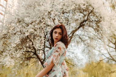 Portrait of charming pretty woman dressed flowery dress posing near apple cherry tree blossoms blooming flowers in the garden park in early spring nature. Fashion, girl model with black hair