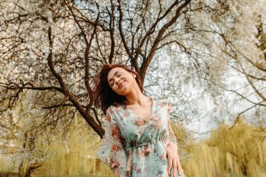 Portrait of charming pretty woman dressed flowery dress posing near apple cherry tree blossoms blooming flowers in the garden park in early spring nature. Fashion, girl model with black hair