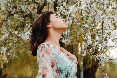 Profile pose looking to side woman dressed flowery dress posing near apple cherry tree blossoms blooming flowers in the garden park in early spring nature. Fashion, girl model with black hair