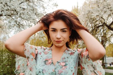 Portrait of charming pretty woman dressed flowery dress posing touching hair near apple cherry tree blossoms blooming flowers in the garden park in early spring nature. Fashion holiday