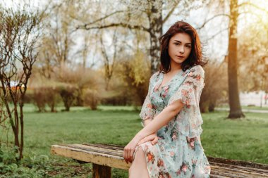 Portrait of charming pretty woman dressed flowery dress sitting on bench near apple cherry tree blossoms blooming flowers in the garden park in early spring nature. Fashion, holiday