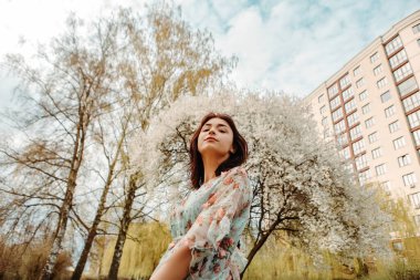 Portrait of charming pretty woman dressed flowery dress posing near apple cherry tree blossoms blooming flowers in the garden park in early spring nature. Fashion, girl model with black hair