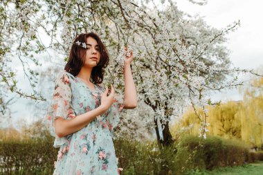 Portrait of charming pretty woman dressed flowery dress posing near apple cherry tree blossoms blooming flowers in the garden park in early spring nature. Fashion, girl model with black hair