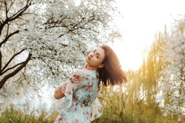 Woman looking over the shoulder dressed flowery dress posing near apple cherry tree blossoms blooming flowers in the garden park in early spring nature. Fashion, girl model with black hair